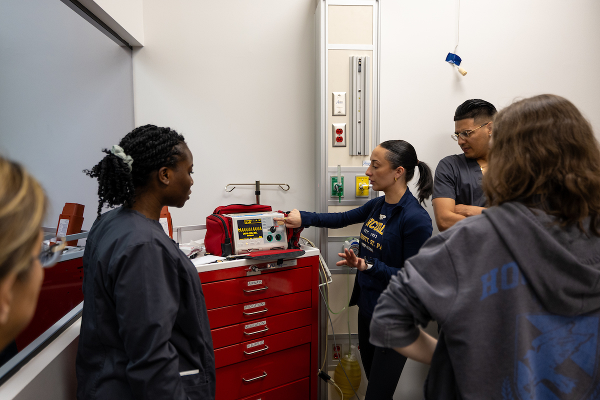 Nursing students standing in a class around a machine that an instructor is pointing at.