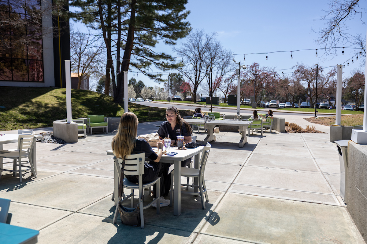Two students sitting outside at a wooden table with snacks on the Denver campus.