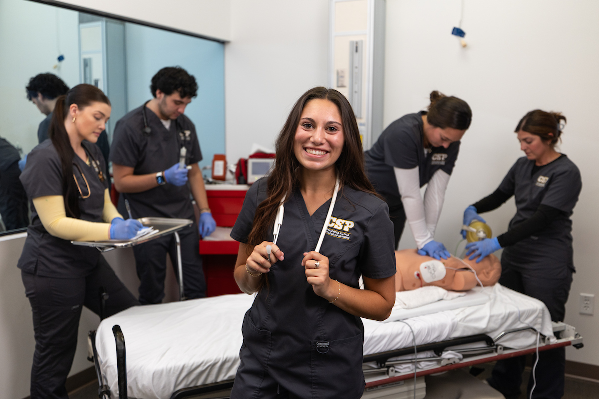 A CSP nursing student in scrubs posing with a stethoscope around her neck and fellow CSP students in the background practicing on a medical manikin.